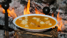 une assiette creuse avec une soupe orangée et des croutons de pain est présentée devant un feu de cheminée allumé